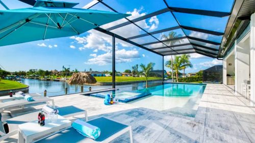 A modern pool area with lounge chairs, an umbrella, and a scenic view of water and palm trees under a clear blue sky.