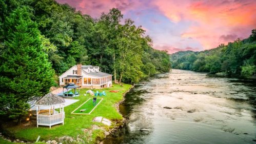 A serene riverside home surrounded by trees, with a gazebo and colorful sunset sky reflecting on the water.