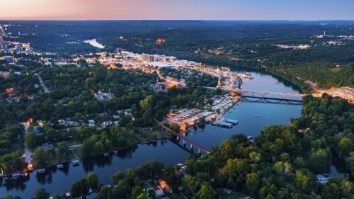 Aerial view of a river town at dusk, with bridges, greenery, and city lights reflecting on the water.