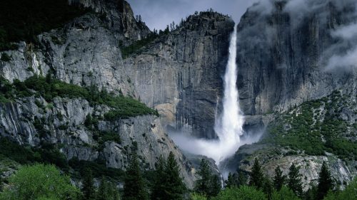 A majestic waterfall cascades down rocky cliffs, surrounded by lush greenery and misty clouds in a dramatic landscape.