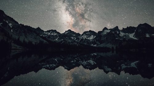 A starry night sky reflects over a calm lake, surrounded by rugged mountains and snow-capped peaks.