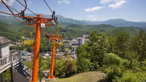 A scenic view from a mountain lift, overlooking a town surrounded by lush green mountains under a clear blue sky.