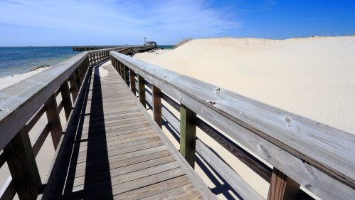 A wooden boardwalk leads to a sandy beach and pier under a clear blue sky.