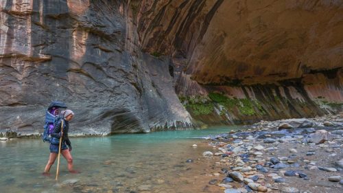 A hiker wades through a river in a narrow canyon, surrounded by towering rock walls and lush greenery.