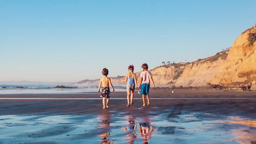 Three children walking along a beach, with cliffs in the background and a clear blue sky above.