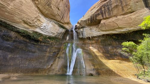 A stunning waterfall cascades between towering sandstone cliffs under a bright blue sky. Lush greenery surrounds the scene.