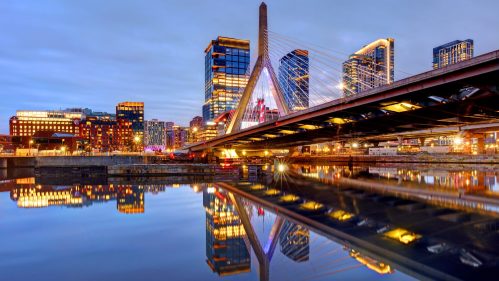 A city skyline at dusk with a bridge reflecting in the water below, illuminated by city lights.
