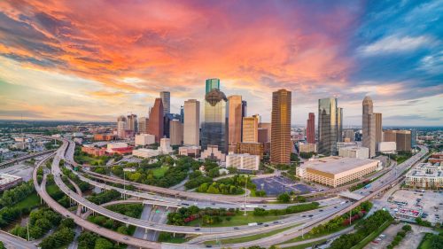Aerial view of Houston's skyline at sunset, featuring skyscrapers and vibrant clouds over highways and greenery.