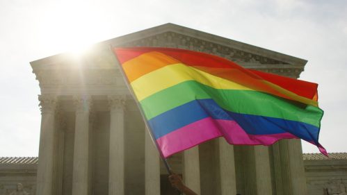 A person holds a rainbow flag in front of a classical building, with sunlight shining in the background.