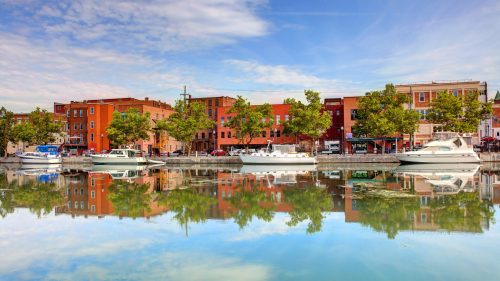 A serene waterfront scene with boats and colorful buildings reflecting in calm water under a blue sky.