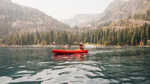 A person paddles a red kayak on a calm lake surrounded by mountains and dense evergreen trees.