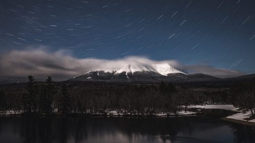 A serene night scene of a snow-capped mountain under star trails, reflected in a calm lake surrounded by trees.