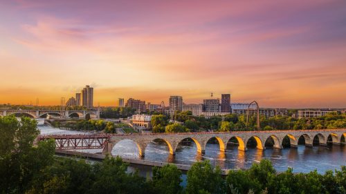 A scenic view of a city skyline at sunset, featuring bridges over a river and lush greenery along the banks.