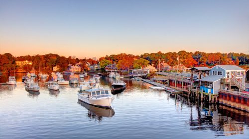 A serene harbor at sunset, featuring boats moored in calm waters and colorful trees lining the shore.