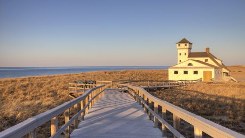 A wooden walkway leads to a white building by the beach, with tall grass and a calm ocean in the background at sunset.