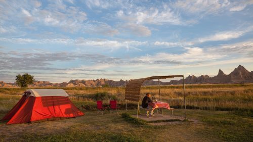 A person sits at a table under a shelter near a red tent, with scenic mountains and a blue sky in the background.