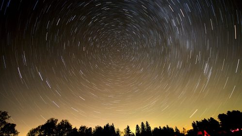A starry night sky with circular star trails around a central point, silhouetted trees in the foreground.