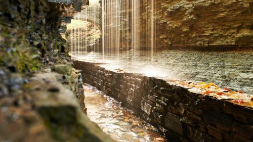 A serene canyon with a narrow stream and cascading waterfalls, surrounded by rocky walls and autumn leaves.