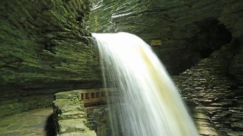 A serene waterfall cascades over moss-covered rocks in a narrow, green-hued gorge.