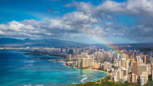 Aerial view of a vibrant city by the ocean, featuring skyscrapers, a rainbow, and lush greenery along the coastline.