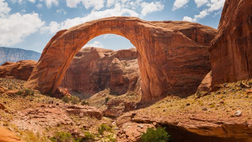Natural rock arch in a desert landscape, surrounded by red rock formations and a blue sky with scattered clouds.