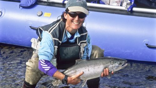 A smiling person in a blue shirt and cap holds a large fish by a river, with a canoe in the background.