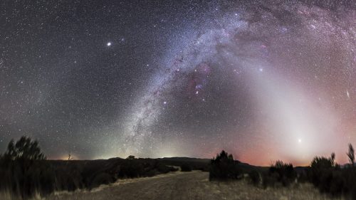 A panoramic view of a starry night sky with the Milky Way, colorful nebulae, and a faint glow on the horizon.