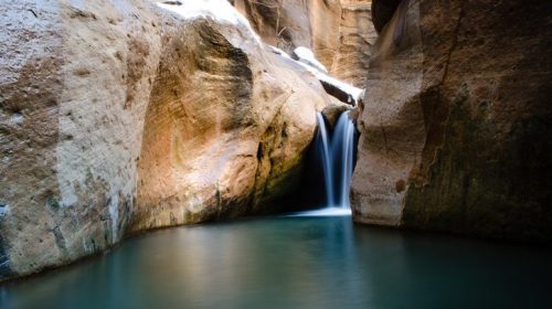 A serene waterfall cascades into a tranquil pool, surrounded by towering rock formations and a hint of snow.