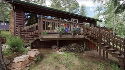 A cozy log cabin with a wooden deck, surrounded by trees and colorful flower pots, set in a natural landscape.