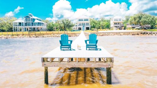 Two blue chairs on a dock overlooking a calm waterway, with houses and a bright blue sky in the background.