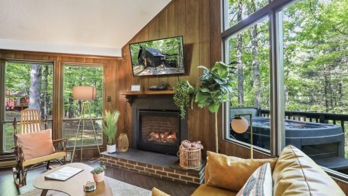Cozy living room with a fireplace, large windows, and a view of trees, featuring a TV and comfortable seating.