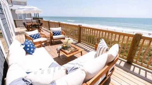 Cozy beachside deck with seating, a table, and ocean views under a clear blue sky.