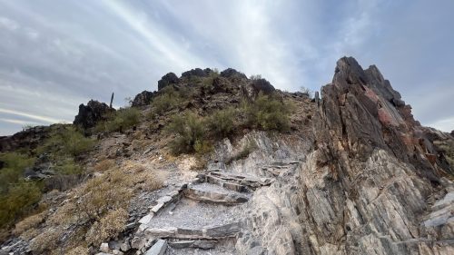 A rocky trail leads up a hillside with sparse vegetation under a cloudy sky.