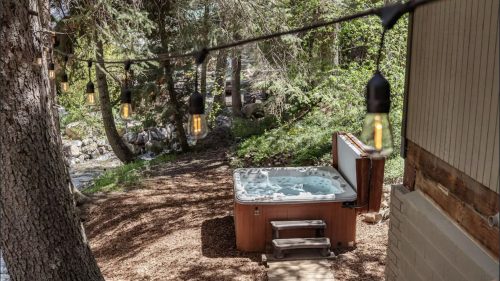 A serene outdoor scene featuring a hot tub surrounded by trees and string lights, near a flowing stream.