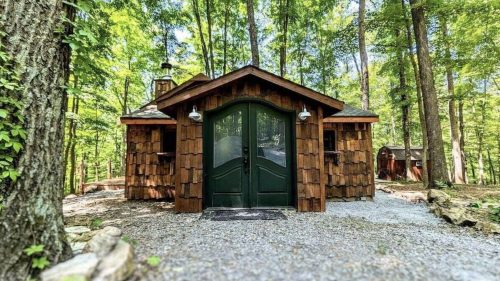 A charming wooden cabin surrounded by lush green trees, featuring a gravel path and large double doors.