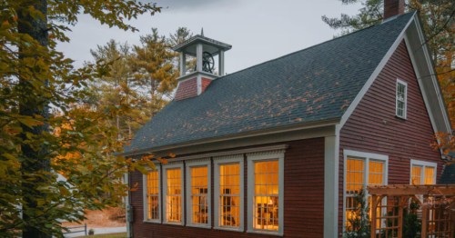 A small red schoolhouse, now converted into an Airbnb, in a field surrounded by fall foliage.