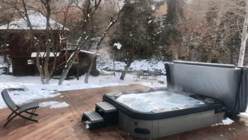 A hot tub on a wooden deck surrounded by snow-covered trees and a cozy cabin in the background.