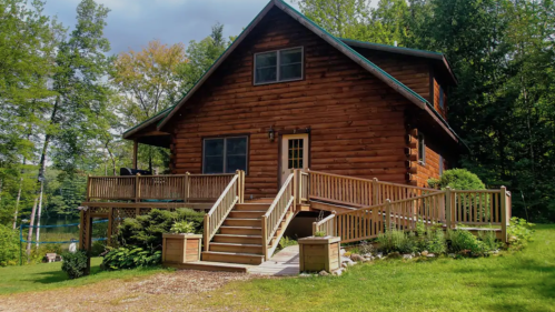 A rustic wooden cabin surrounded by trees, featuring a front porch and landscaped yard.