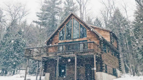 A wooden cabin surrounded by snow-covered trees, with large windows and a balcony, in a winter landscape.