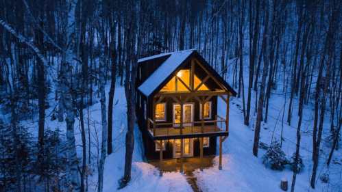 A cozy cabin in a snowy forest, illuminated warmly at dusk, surrounded by tall trees.