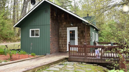 A cozy green cabin with wooden accents, surrounded by trees and a small deck leading to a stone pathway.
