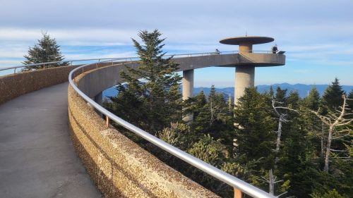 A curved observation deck surrounded by trees, with a circular viewing platform at the end, under a blue sky.