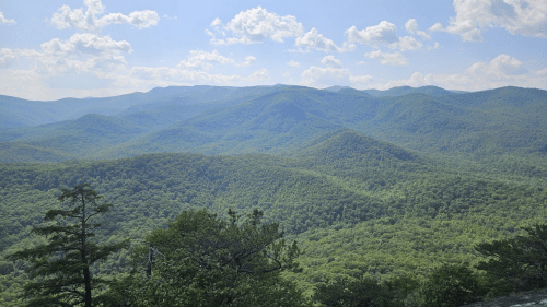 A panoramic view of lush green mountains under a bright blue sky with scattered clouds.