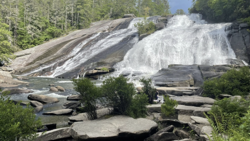 A cascading waterfall flows over rocky terrain, surrounded by lush greenery and trees under a partly cloudy sky.