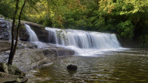 A serene waterfall cascades over rocks into a calm pool, surrounded by lush green trees and foliage.