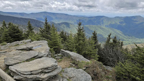 A rocky outcrop overlooks a lush green valley and distant mountains under a cloudy sky.