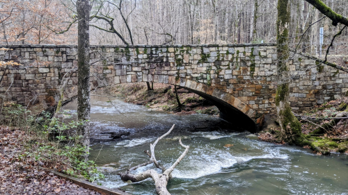 A stone arch bridge over a flowing creek, surrounded by trees and a forested landscape.