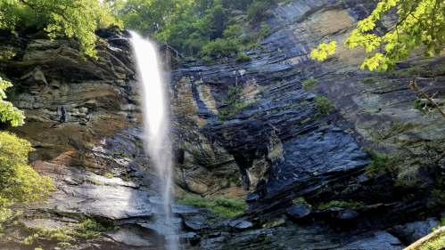 A tall waterfall cascades down rocky cliffs, surrounded by lush greenery and a serene natural setting.