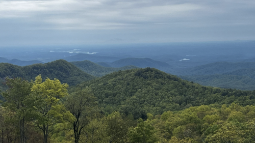 A panoramic view of rolling green mountains under a cloudy sky, with a distant lake visible in the valley.