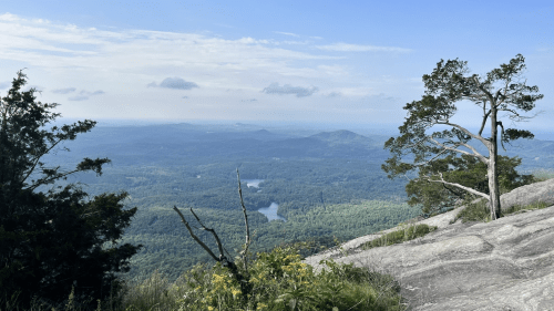 A panoramic view from a rocky outcrop, showcasing lush green valleys, distant mountains, and a serene lake under a blue sky.
